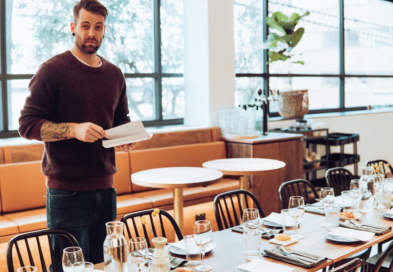 Man setting table with glassware and silverware in sunlit modern restaurant interior.