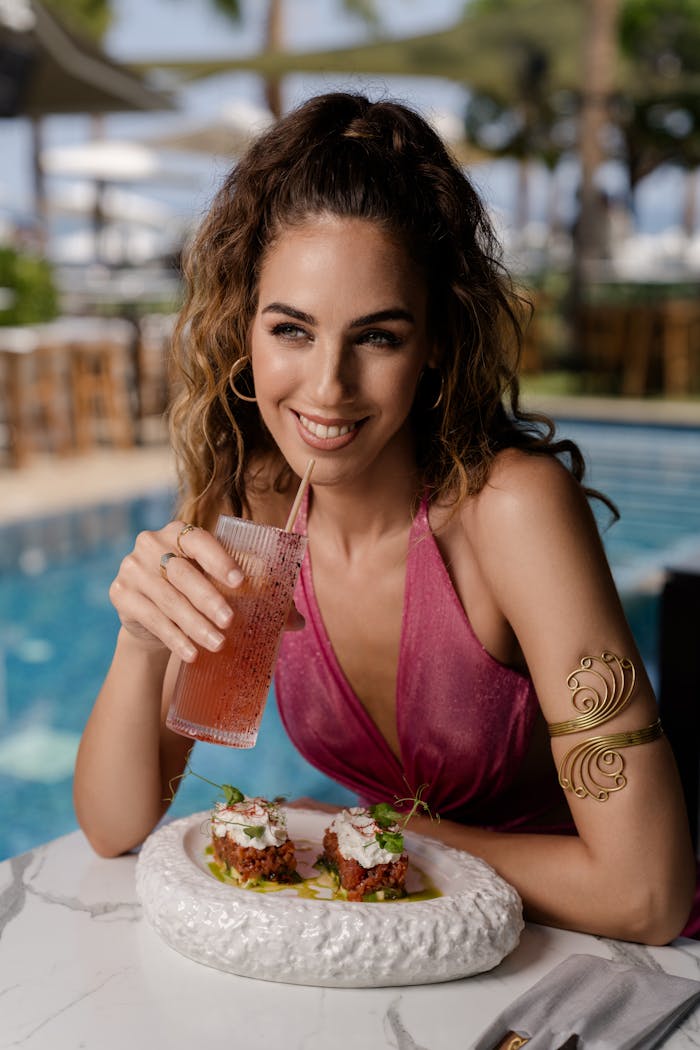Young woman in pink dress enjoying a refreshing drink and meal by the poolside on a sunny day.